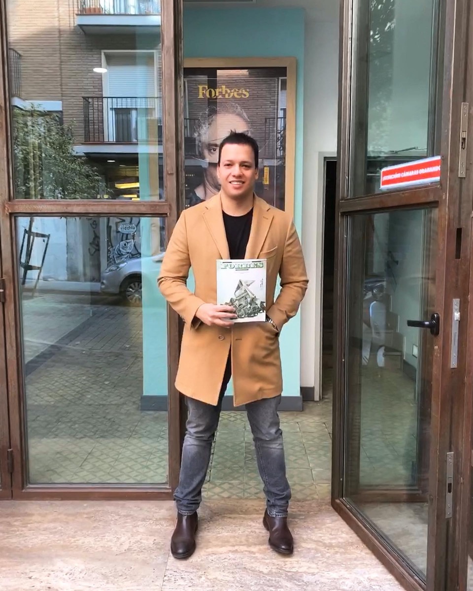 Joshua con abrigo camel sosteniendo una revista Forbes en la entrada de la revista