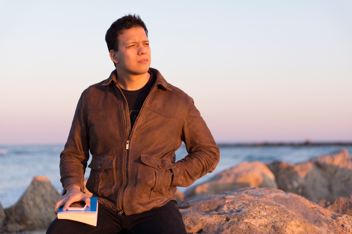 Joshua sentado en las rocas junto al mar al atardecer, con un libro en la mano