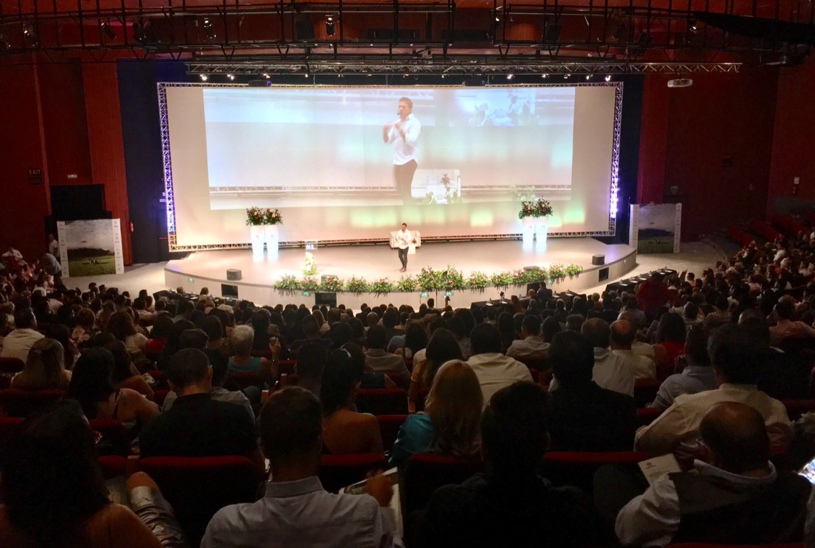 Joshua speaking on a large stage to a full auditorium, projected on the screen behind him.